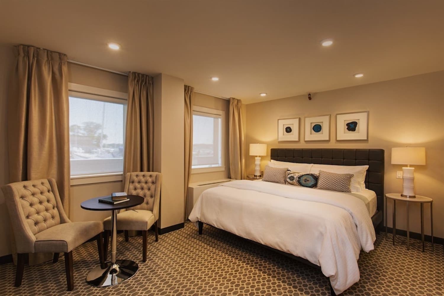 A bedroom with carpeted floor, a king bed with white bedding, night stands with lamps on either side of the bed, and a sitting area with a plush chairs and a small table.