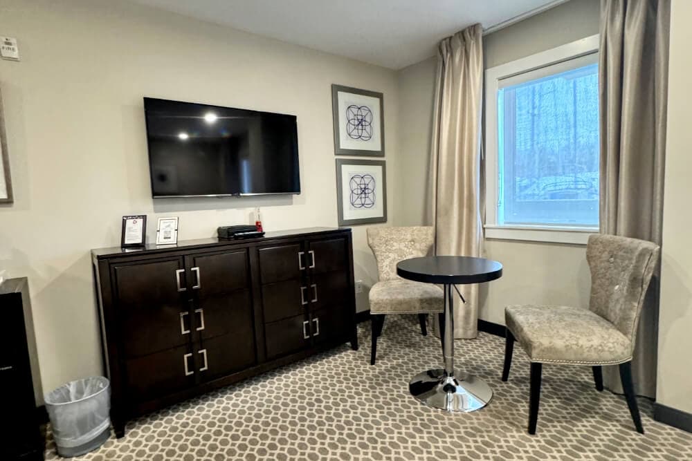 A corner area in a room with a dresser, flat screen TV and 2 plush chairs with a small table.
