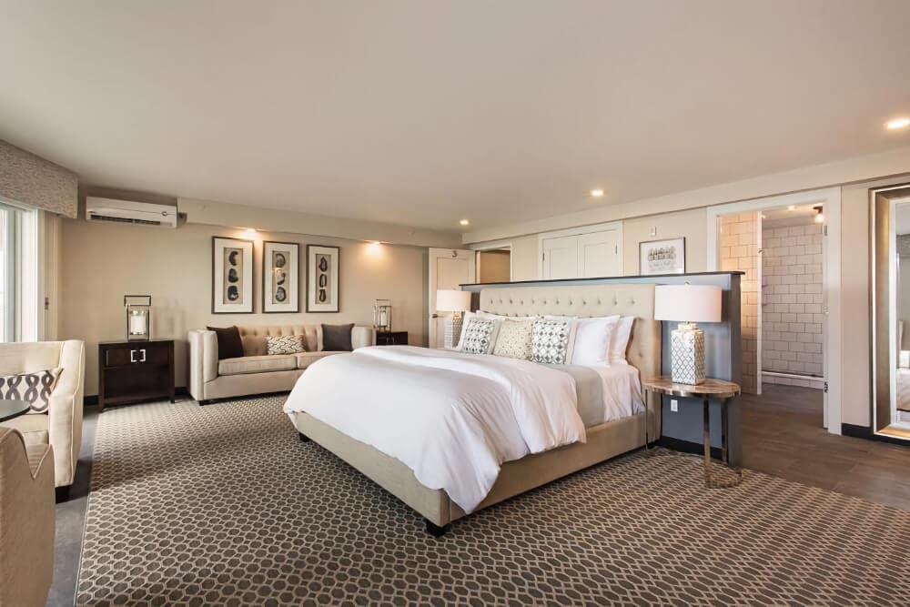 A spacious bedroom with a king bed, side tables with lamps on either side, a sofa and separate sitting area with table and chairs.