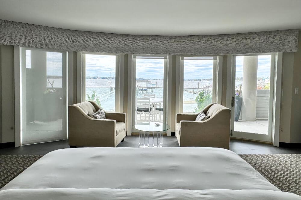 A bedroom with part of a bed, and 2 oversized plush armchairs facing each other with a small table in between them, a bank glass doors that open out to a patio with views of a marina.