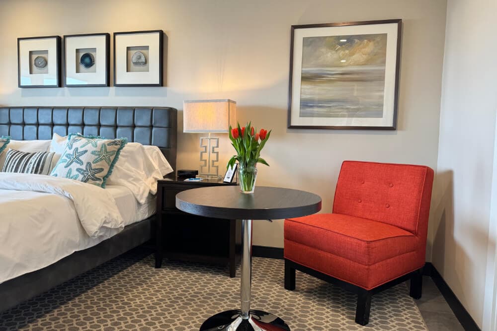 A bedroom with a bed with white bedding and blue starfish throw pillows, a table with red flowers in a vase, and a plush red chair.