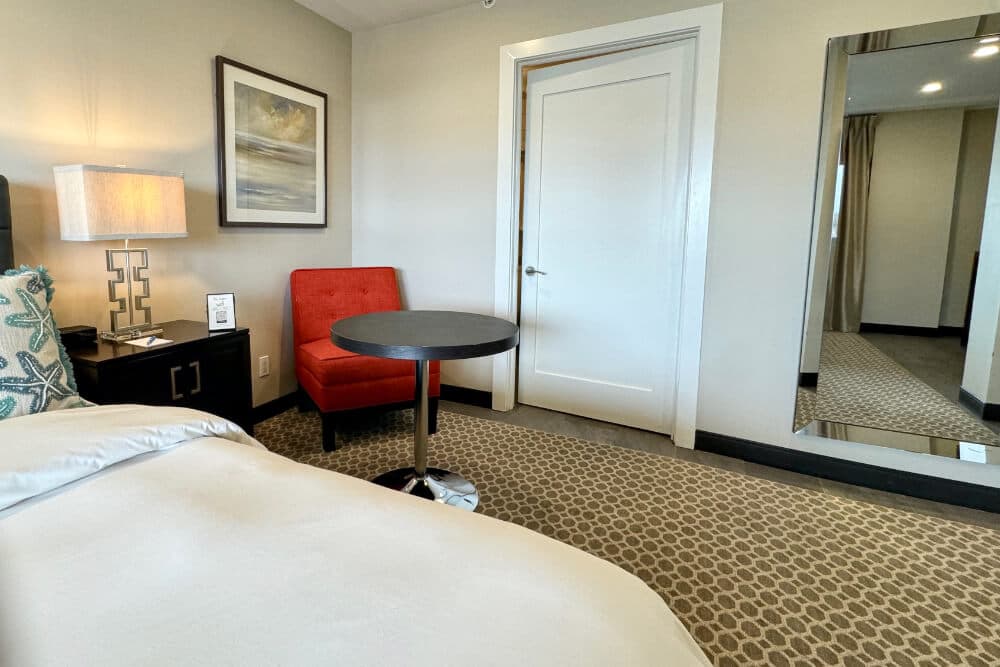 A bedroom with a bed with white bedding and blue starfish throw pillows, a table with red flowers in a vase, and a plush red chair.