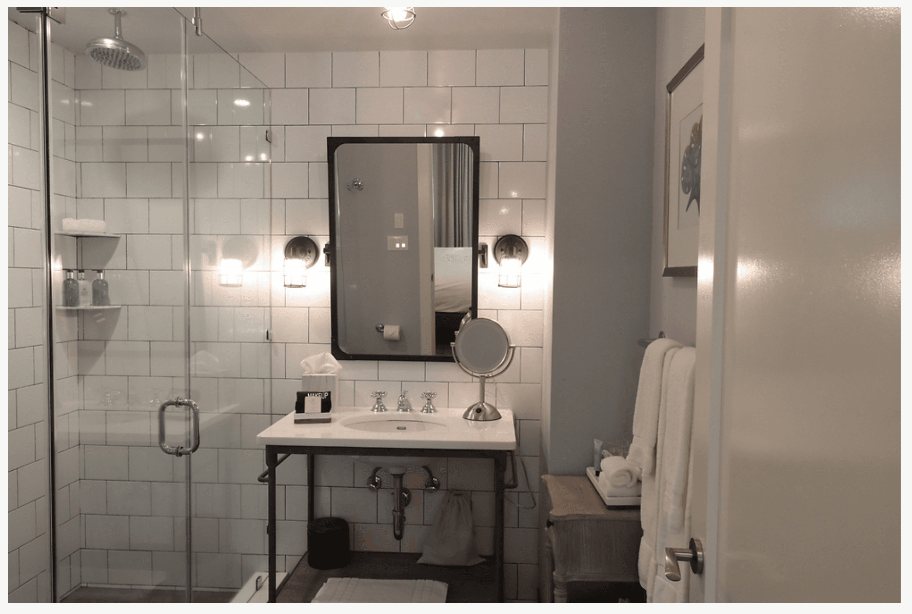 A chic bathroom with a walk in shower with clear glass sides, a vanity with sink and makeup mirror