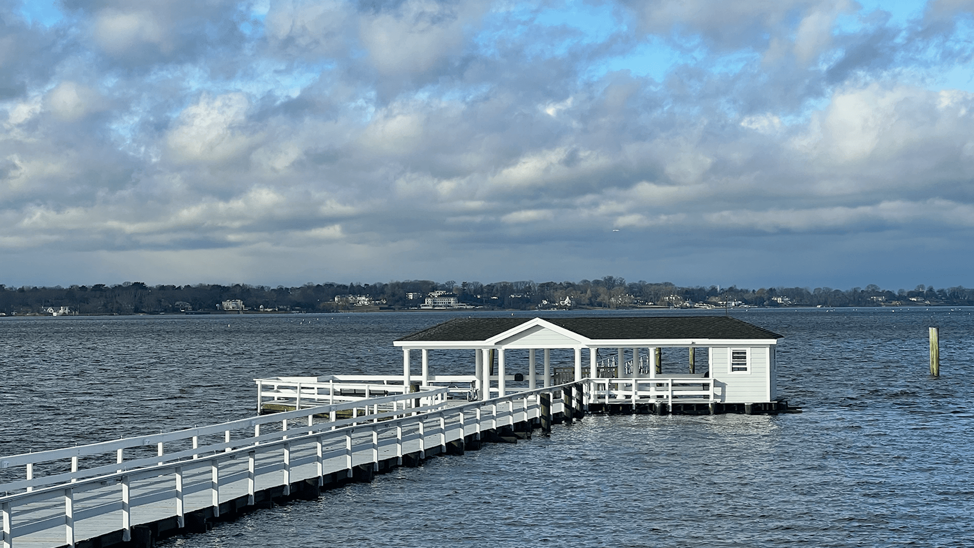 A white dock extends into a calm body of water under a partly cloudy sky.