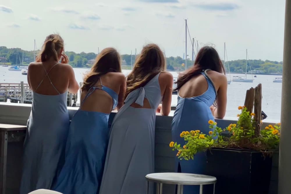 bridesmaids in blue formal dresses leaning over a railing looking out onto the water