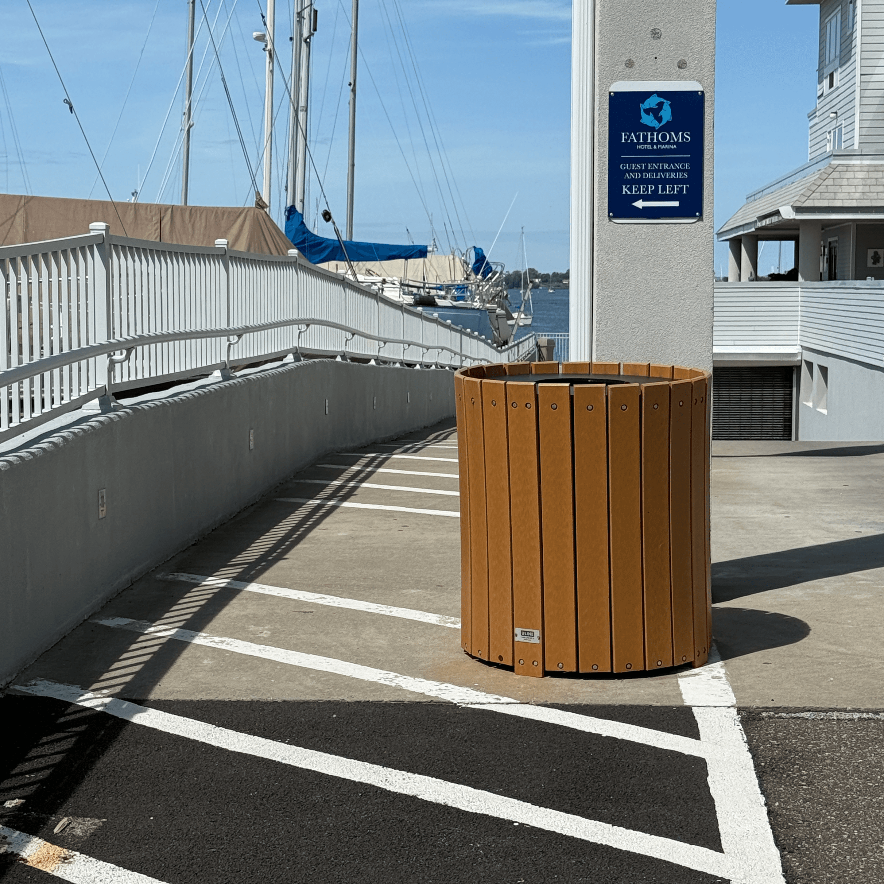A wooden trash bin is positioned beside a waterfront walkway with sailboats in the background.