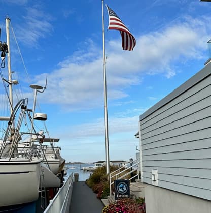 A walkway with boats tied up to a dock on one side, and a flagpole with an American Flag on the other side by a sign that says Fathoms.
